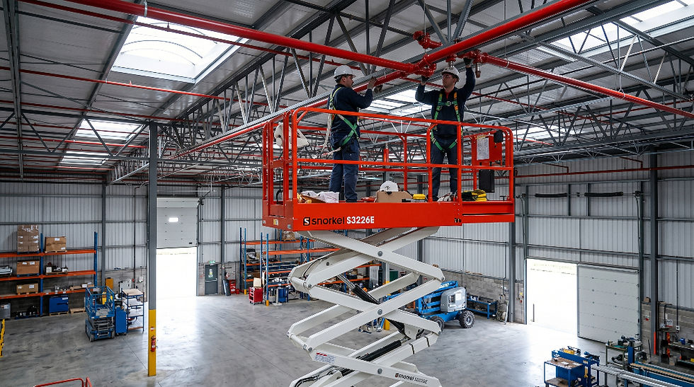 Dos trabajadores con cascos en una plataforma elevadora naranja, instalando tuberías rojas en un almacén industrial amplio y bien iluminado.