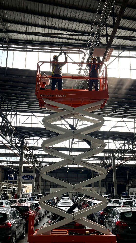 Trabajadores en una plataforma elevadora dentro de una fábrica de autos. Estructura metálica, luz natural. Cartel de Ford al fondo.