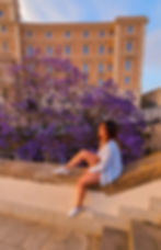 Portrait en pied de Manon Laterza, assise sur un muret au coucher de soleil, devant un arbre fleuri violet à Cagliari (Sardaigne)