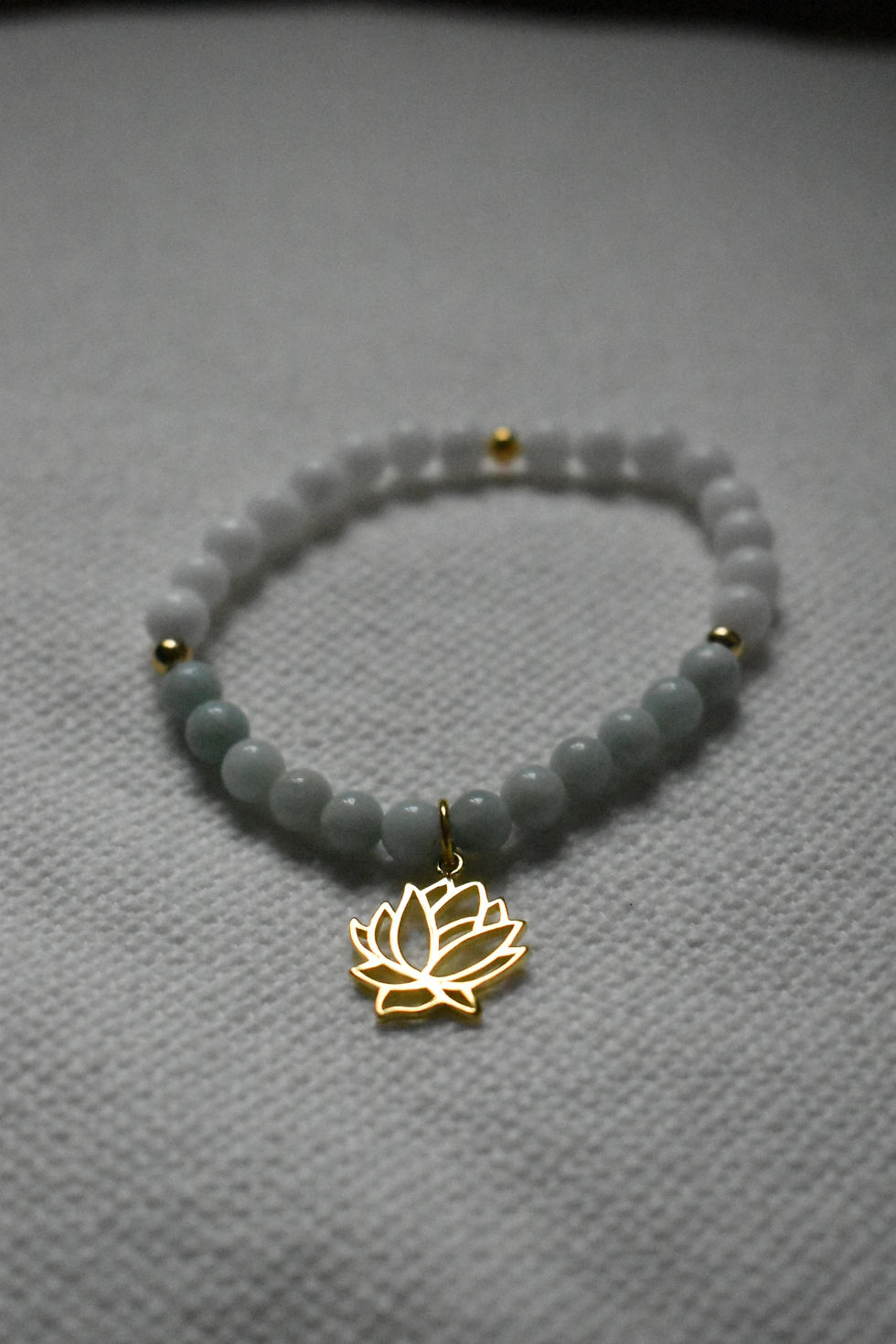 Thumbnail: Armband mit weissen und hellblauen Perlen und einem goldfarbenen Lotusblütenanhänger. Das Armband ist auf einem hellen Untergrund