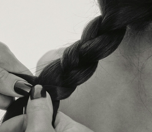 A black and white image shows a woman braiding her hair while seated in a hair salon.