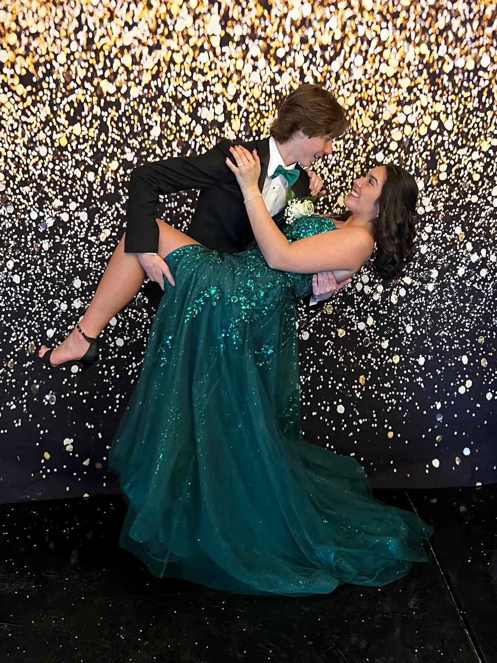 A guy in a black tuxedo lifts a girl in a green sparkly dress at prom. They smile at each other against a glittery gold and black backdrop.
