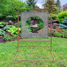 Garden art with hanging antique floor grate filled with marbles set in resin and hanging basket in centre. Hanging basket is a hypertufa planter sitting in a rusty brake rotor