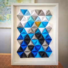 Art created with origami folded pyramids in shades of blue, black, grey, white and one metallic gold. Displayed in frame with plexiglass cover.