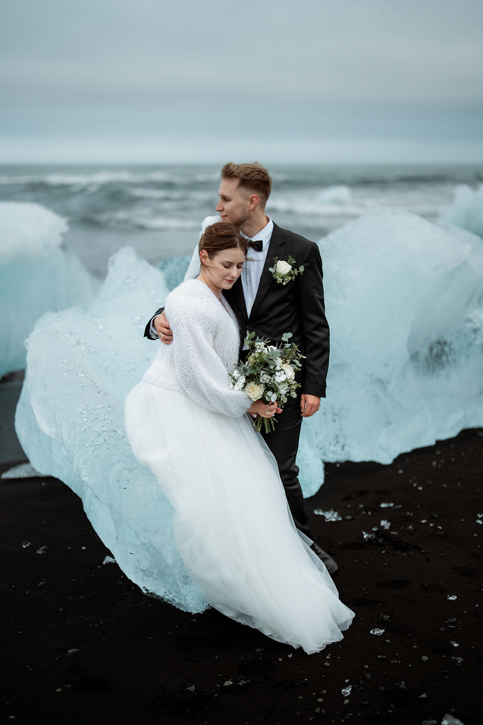 bride and groom embrace on a black sand beach, surrounded by icebergs. Overcast sky, bride in white, groom in black suit, holding flowers during iceland elopement