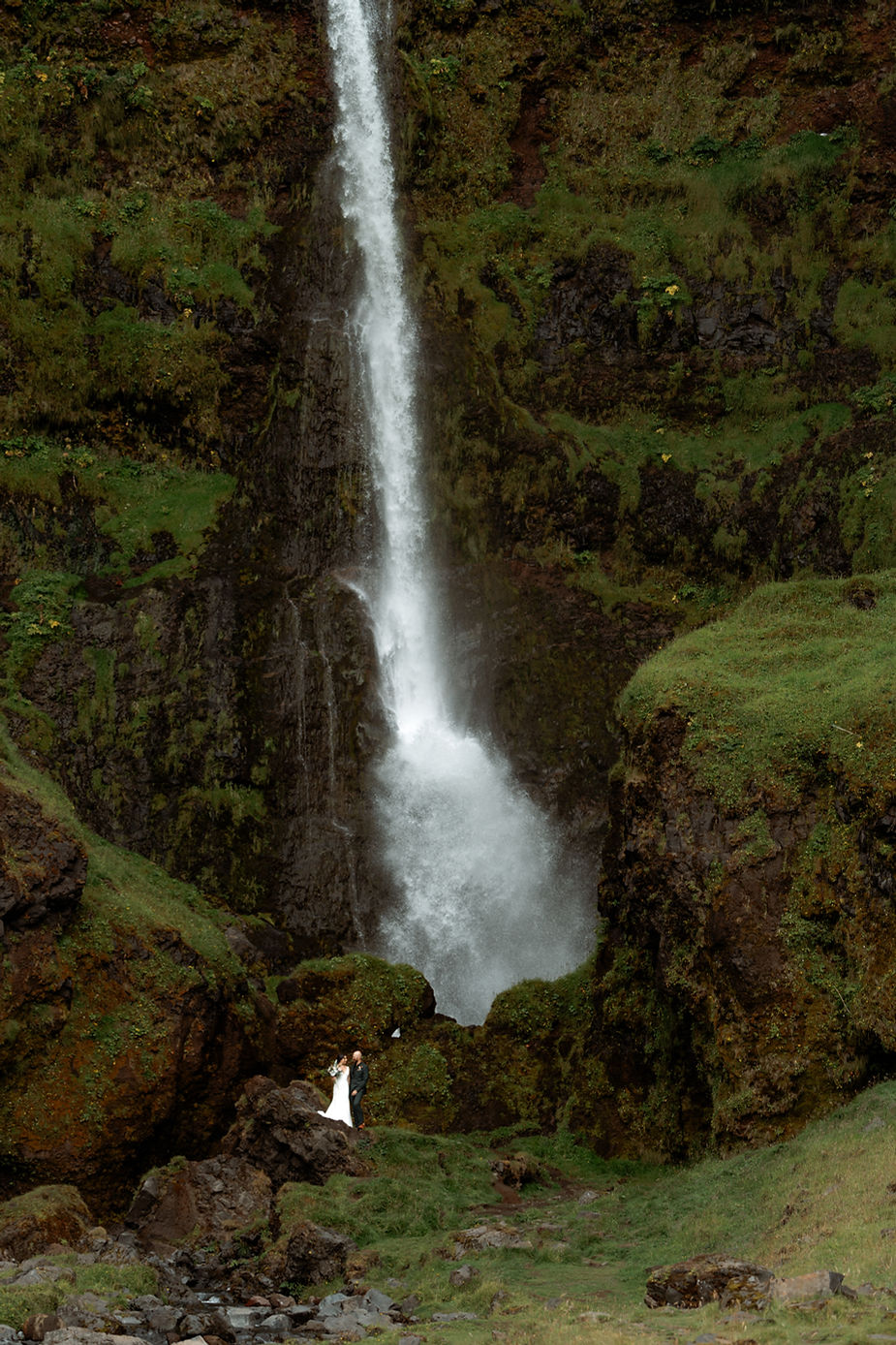 Bride and groom stand in the distance on grassy area at the base of a waterfall during elopement.