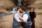 Two grooms in blue vests kiss outdoors, with a waterfall in the background. An officiant wearing a brown robe smiles nearby during Ásatrú elopement ceremony