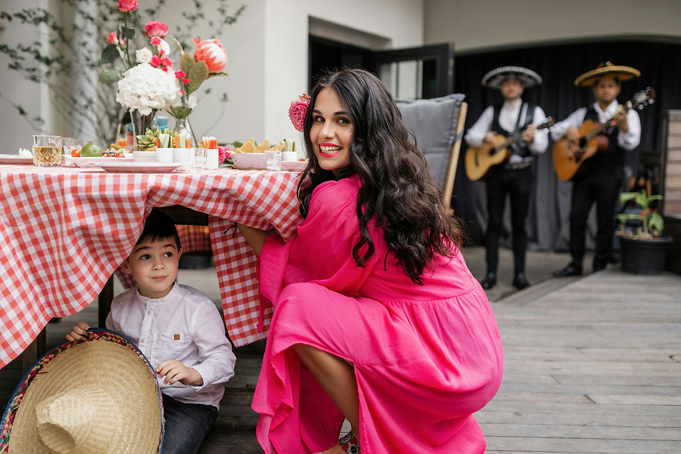 Woman in pink dress smiles beside a child hiding under a red-checkered table.