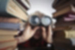 Person in plaid shirt peers through binoculars between stacks of old books.