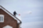 Person getting snow off a brick roof with a red shovel. Overcast sky, snow flying in mid-air.
