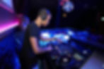 DJ in a dimly lit club mixes tracks on a console. Neon lights glow in pink and blue.