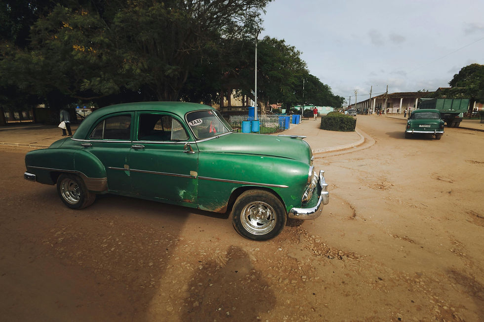 A vintage green taxi with "Taxi" on the windshield is parked on a dirt road near trees and blue barrels, under a cloudy sky.