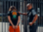 A police officer detains a person in handcuffs against a metal fence. The detainee wears a black shirt with colorful design and text "KushKilla."