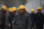 Construction workers in yellow helmets walk on a dusty site; focus on a man in a dark jacket