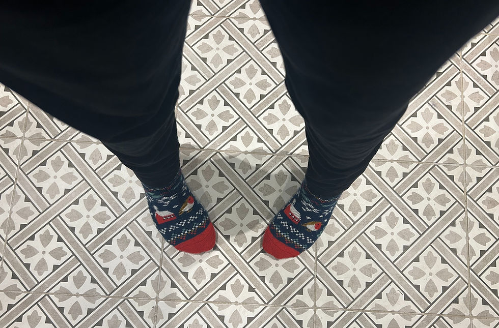 Feet in festive socks with snowman and bird design on tiled floor with geometric pattern. Dark pants visible. Cozy and playful mood.