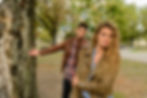 Woman in brown jacket looks serious, arms crossed. Man in background gestures behind a tree.