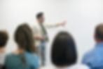 Man giving a presentation with a tablet, gesturing at a blank whiteboard. Audience in the foreground.