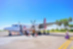 Small plane on runway under clear blue sky, passengers boarding with luggage. Palm trees line the background. Bright, sunny day.