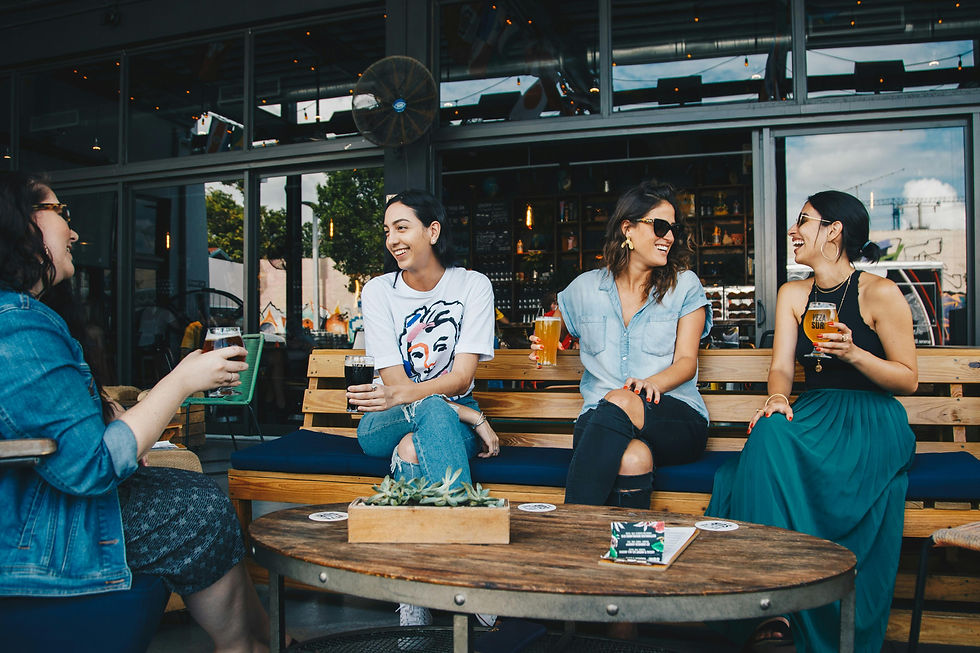 Four women sit outdoors at a bar, laughing and holding drinks.