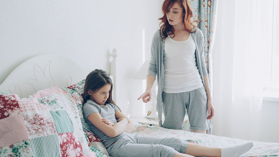 Mother points at an upset girl sitting on a bed in a bright bedroom with floral quilt and white curtains.