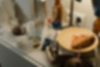 Cluttered table with empty glasses, a wine glass, used tissues, and a pizza slice on a plate, with a lamp in a dimly lit room.