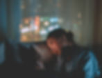 Woman in dim room looks contemplative, holding a smartphone. Blurred city lights visible through window.