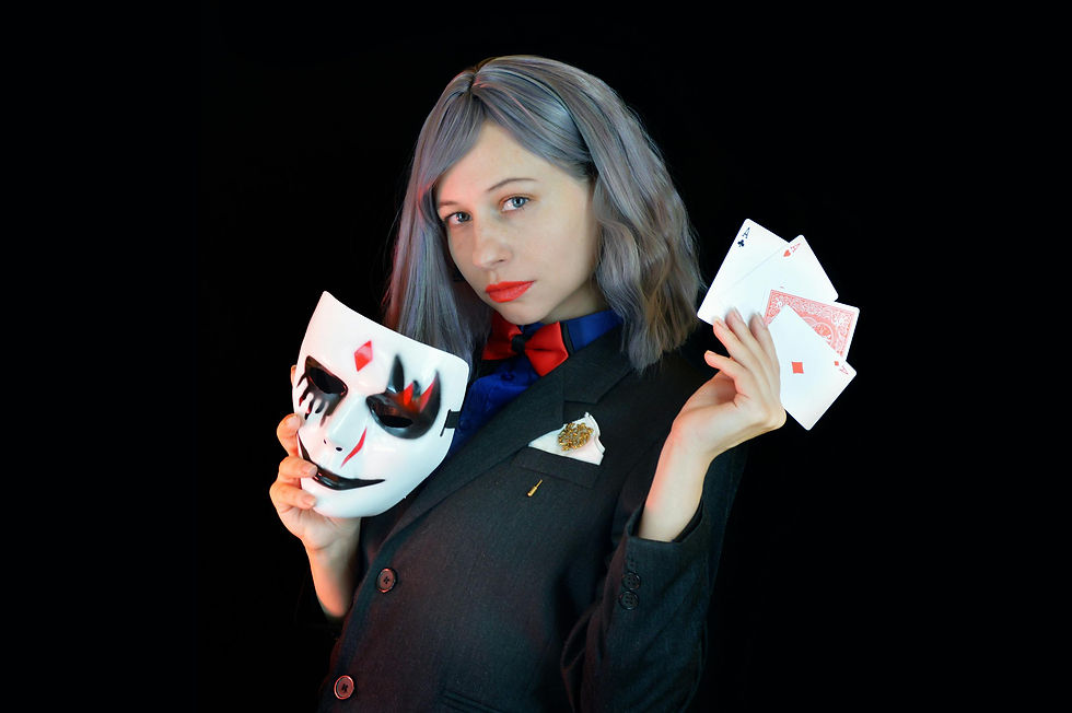Person in a suit holds a painted mask and playing cards against a dark background.