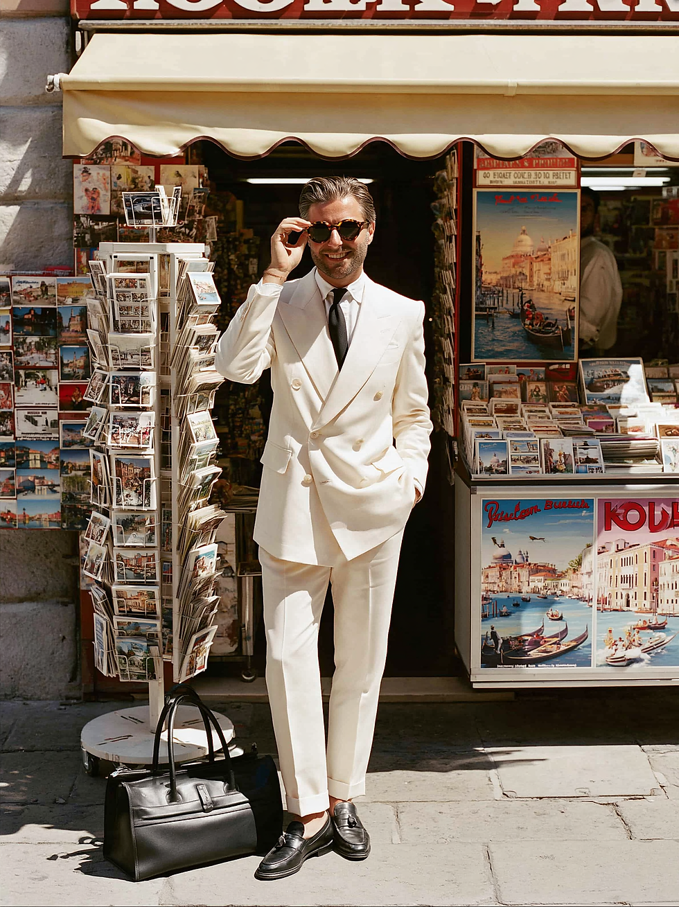 Smiling man in a white suit adjusts sunglasses outside a postcard shop, beside a black bag and racks of travel postcards.
