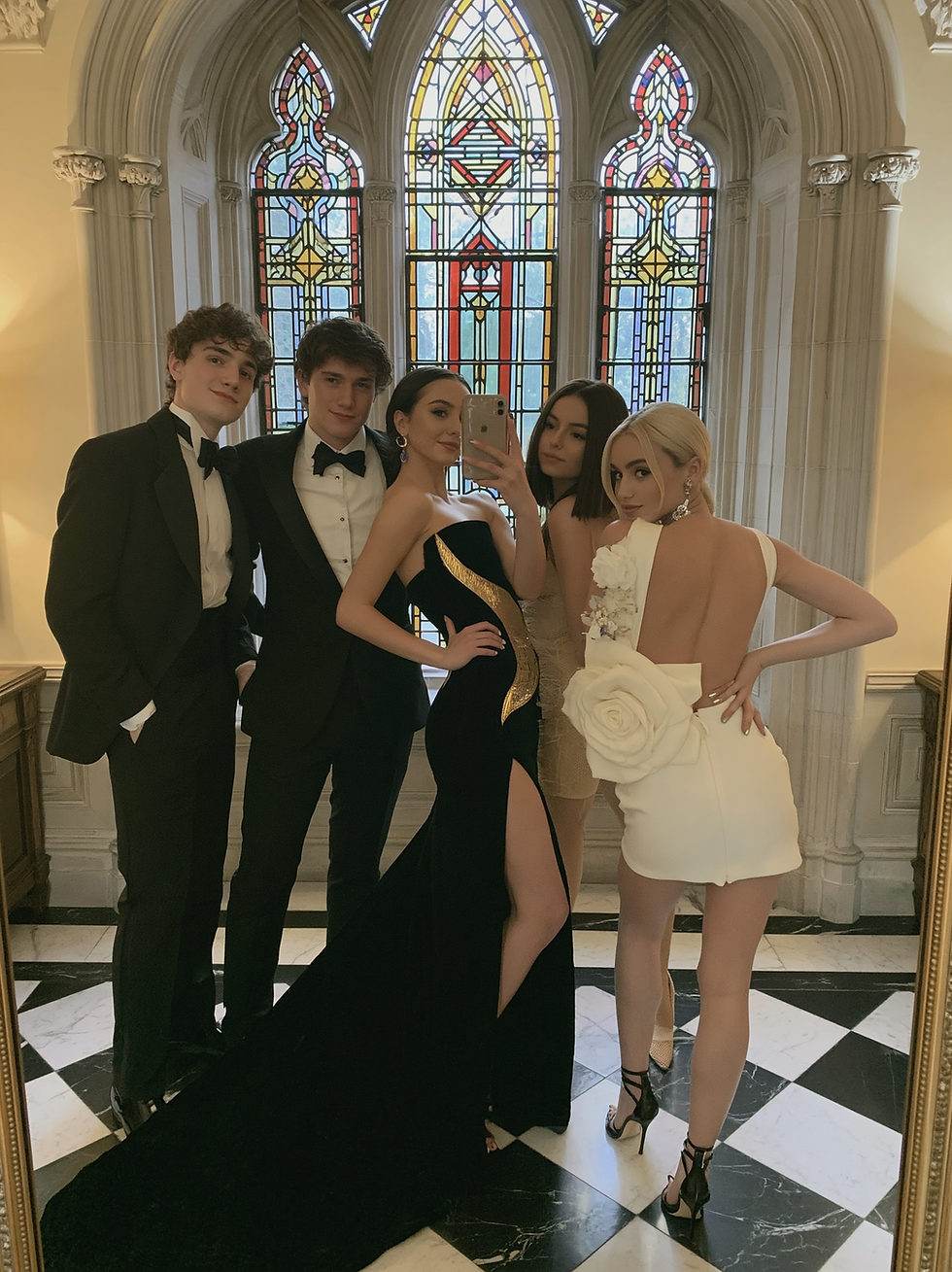 Five young adults pose for a mirror selfie in formal gowns and tuxedos before stained-glass windows in a grand hall