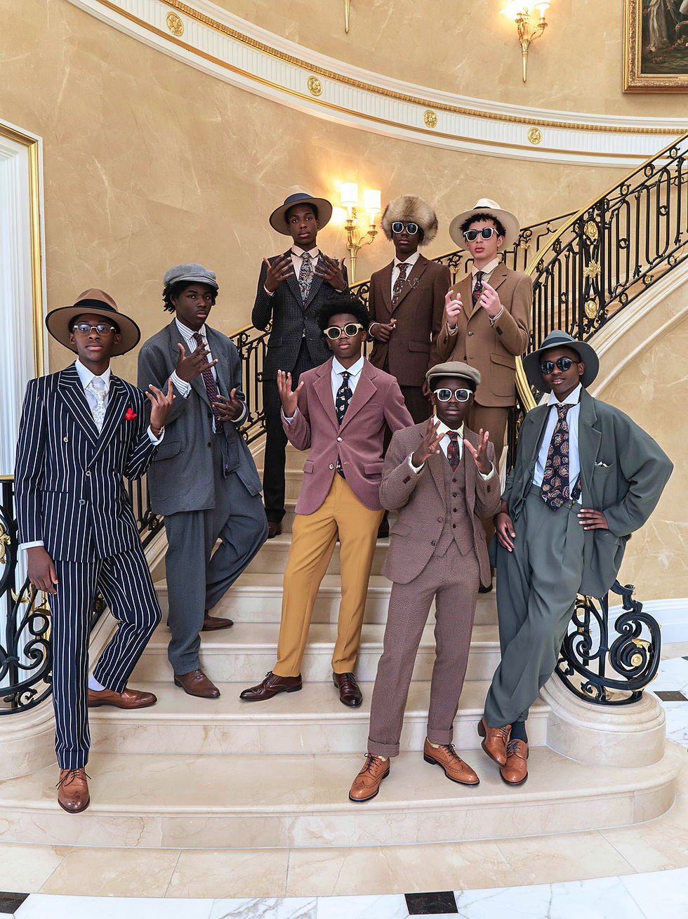 Seven stylish young men in suits and hats pose on a grand marble staircase, wearing sunglasses in an elegant mansion lobby.