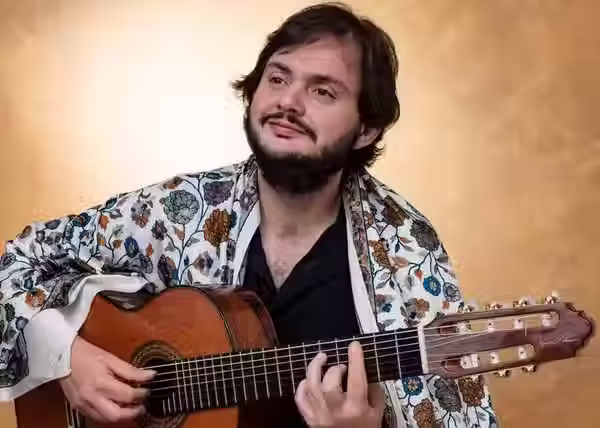 homem, cabelos castanhos, barba curta e bigode com camisa preta e casaco branco com estampa florida verde e dourada tocando violão