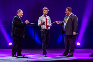 Magician Ryan Joyce on stage with two convention attendees holding a rope