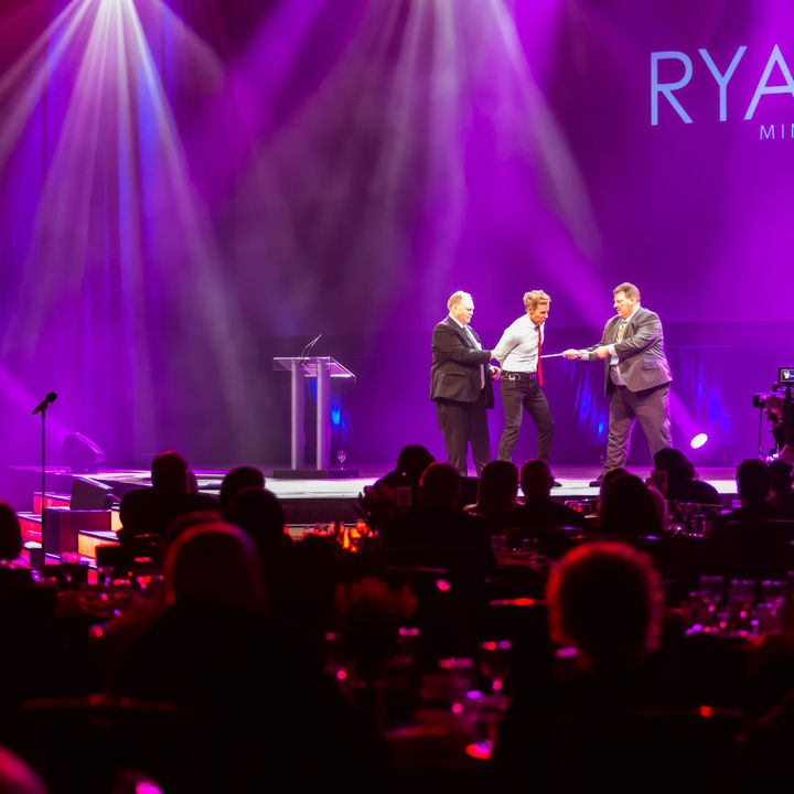 Ryan Joyce performing on a purple-lit stage with two corporate guests, showcasing a moment of clean, interactive comedy.