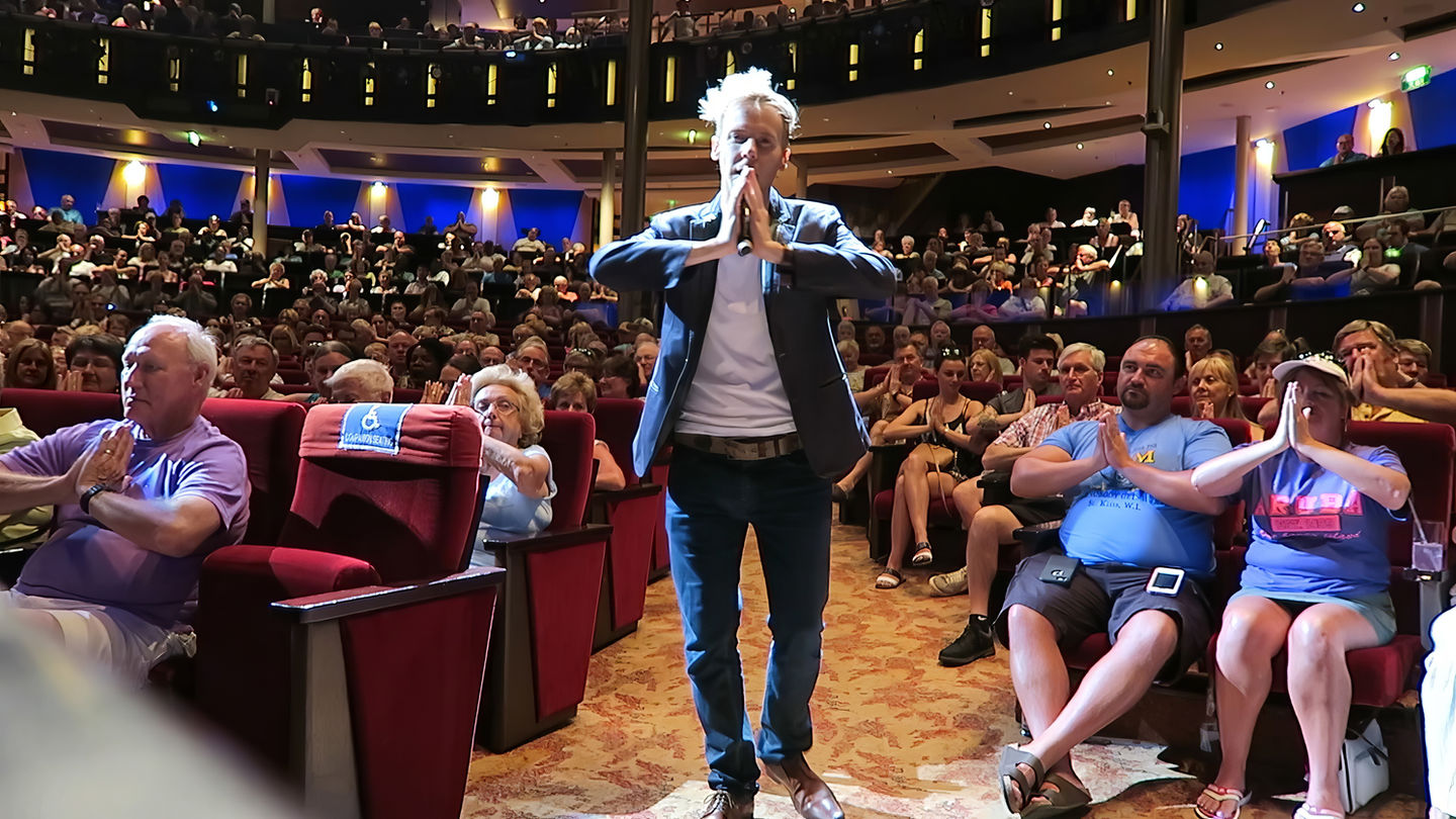 Comedy hypnotist Ryan David on stage in a grey suit, gesturing toward a large, cheering theater audience.