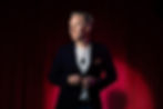 Magician Ryan Joyce in a dark blazer and white shirt stands confidently against a red curtain background. He's adjusting his jacket with a thoughtful look.