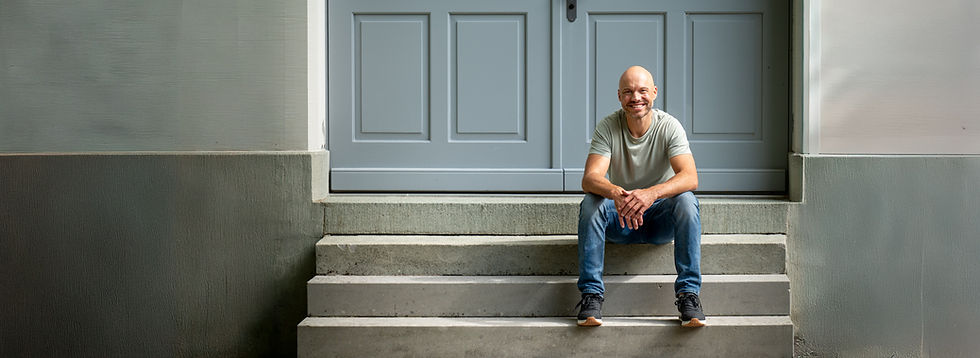 Franz Manser in grauem T-Shirt und blauen Jeans sitzt auf einer Treppe vor einer hellgrauen Tür mit vier rechteckigen Feldern.