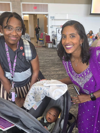 Priya poses with a woman. Between them is a baby in a stroller. 