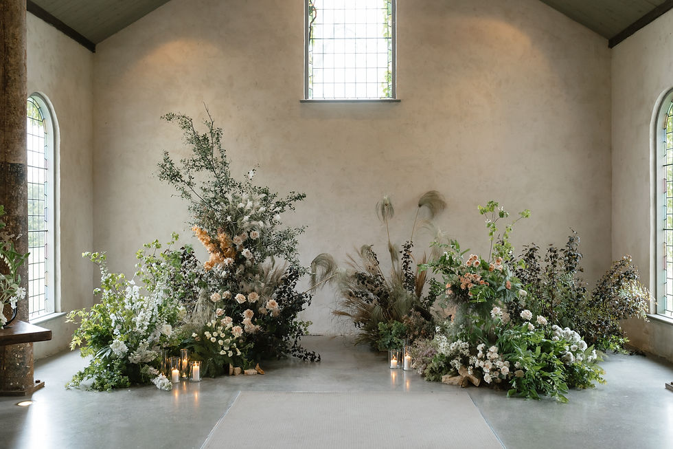 A simple, elegant church interior with large floral arrangements consisting of various flowers and greenery placed on the floor. Two candle clusters are lit at the base of the arrangements.