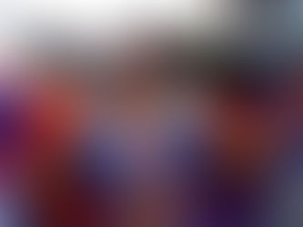 Woman in vibrant blue carnival costume with colorful feathered wings, smiling, holding her hair. Background shows houses and trees.