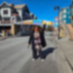 Woman in a black coat stands on a street holding a "2026 Sundance Film Festival" banner. Sunny day, buildings in background.