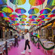 Woman smiling on a vibrant pink street with colorful umbrellas overhead.