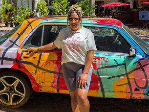 Woman poses smiling beside a colorful, graffiti-covered car. She's wearing a white shirt and gray shorts. Sunlit street and green plants in background.