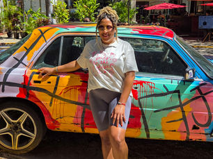 Woman poses smiling beside a colorful, graffiti-covered car. She's wearing a white shirt and gray shorts. Sunlit street and green plants in background.
