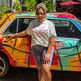 Woman poses smiling beside a colorful, graffiti-covered car. She's wearing a white shirt and gray shorts. Sunlit street and green plants in background.