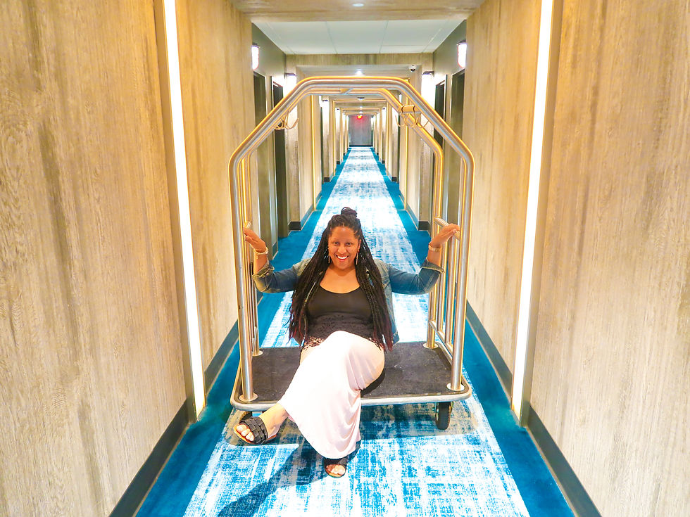 Woman sitting on a luggage cart in a hotel hallway with blue carpet. She is smiling and wearing a denim jacket, creating a joyful mood.