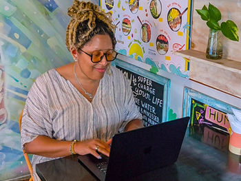 Woman with braided hair and glasses typing on a laptop at a café. Colorful menu and plant in the background. Relaxed and focused mood.