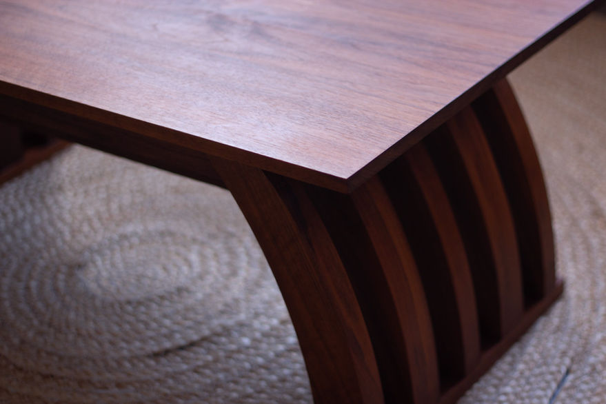 A close-up of a dark brown low-rise table with arched legs is depicted, on a straw-colored woven rug. The focus of the image is of the table top and the arched legs on one side, from an isometric angle.