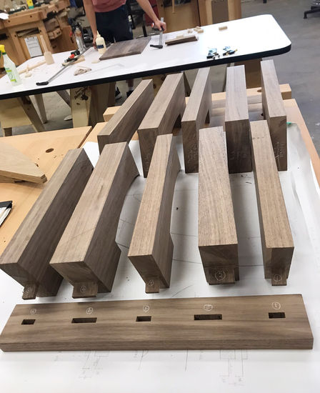 On a work bench, ten table legs are laid out in two rows along with the two rectangular pieces that will connect the legs. Mortises are shown already cut in the connecting pieces, and all the table legs have corresponding tenons.