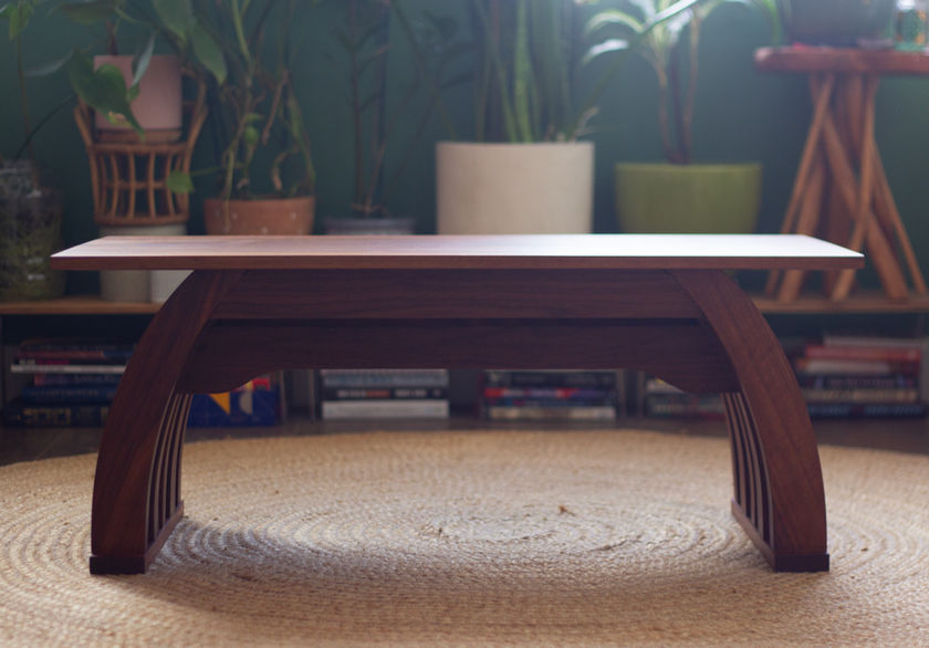 A dark brown low-rise table is depicted from the perspective of front view. Curved legs on both left and right sides arch inward toward the top. The table rests on a straw-colored woven rug, with houseplants and books behind it against a dark green wall.