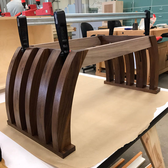 The dry fitting of the entire table sans the table top is depicted on top of a low-rise melamine work surface, with a large piece of craft paper between the piece and the table. The table legs are dry fitted with the aprons via two long clamps running lengthwise and applying clamping pressure between the side aprons attached to the table legs. The table legs and their connectors have finish applied to them already but the front and back aprons are still unfinished.